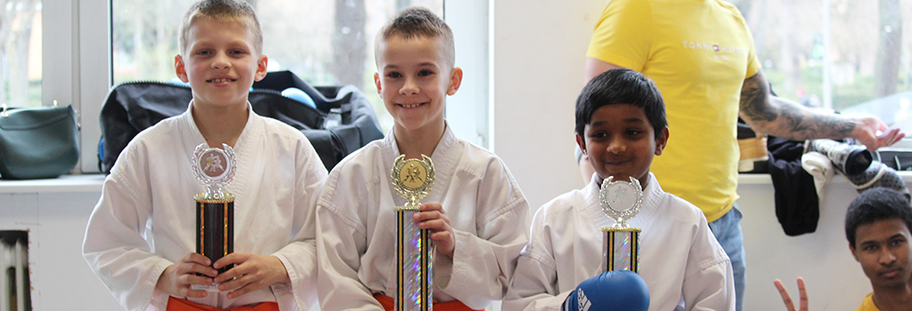 Das Foto zeigt drei stolze Kinder des Karate Club Wedding, die auf dem 2. Oster-Randori '23 ihre verdienten Pokale in den Händen halten. Die strahlenden Gesichter der Kinder zeigen ihre Freude und Begeisterung für den Karatesport und das erfolgreiche Abschneiden beim Turnier.