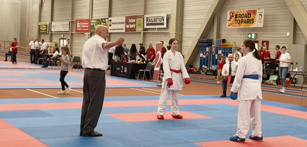 Eine junge weibliche Karateka in weißer Kampfausrüstung steht in einer Kampfposition bei dem Wado-Cup 2024. Der Schiedsrichter neben ihr hebt die Hand, um ihr einen Punkt für eine erfolgreich ausgeführte Karate-Technik zu vergeben. Sie wirkt fokussiert und bereit für die nächste Aktion.